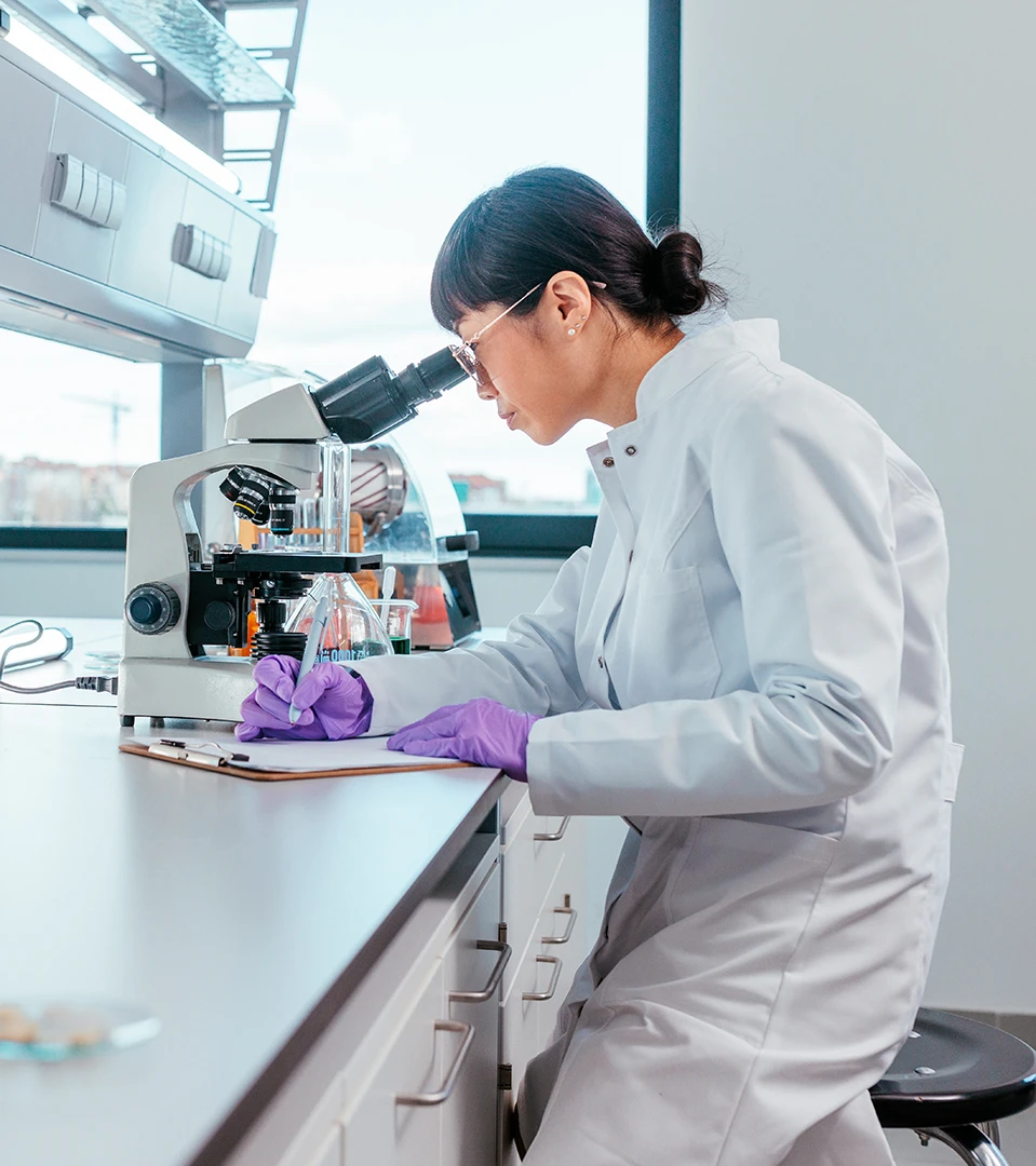 Female scientist sitting at a desk looks at a sample through a microscope